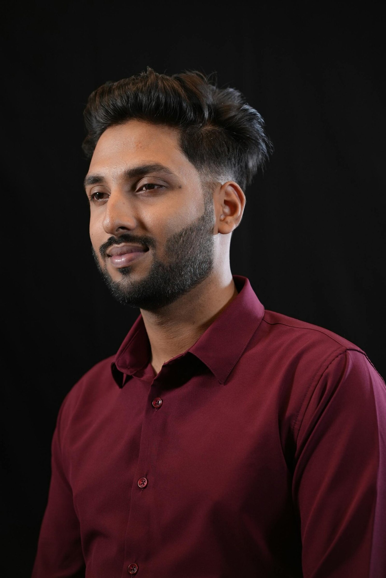 About Professional studio portrait of a young man with a neatly styled beard and maroon shirt against a dark backdrop.